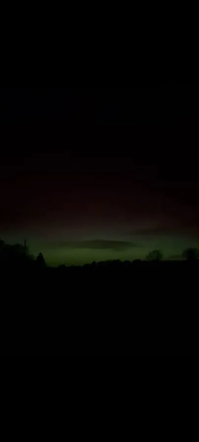 The Northern Lights glowing green and red over the tree line above the field